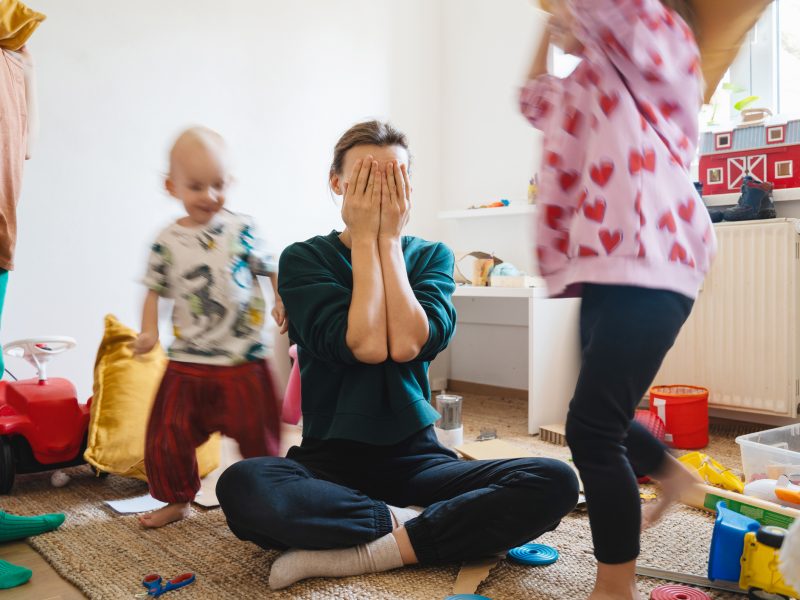 Stressed out mother sitting on floor in middle of toys while children naughty running around her at room. Woman alone burnout with kids. Family home with chaos, mess. Motion blur for speed, real life.